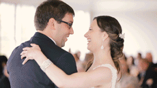 During their first dance at an indoor ballroom reception, a groom lifts his bride as guests look on and clap and cheer