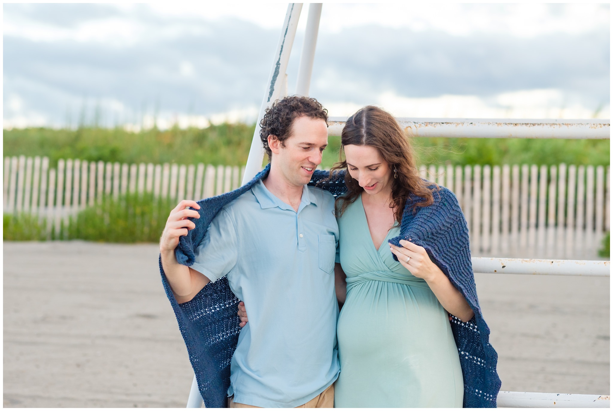 Maternity portraits at Nahant beach at sunset with man in blue and pregnant woman in a green dress wrapped in a crocheted blanket