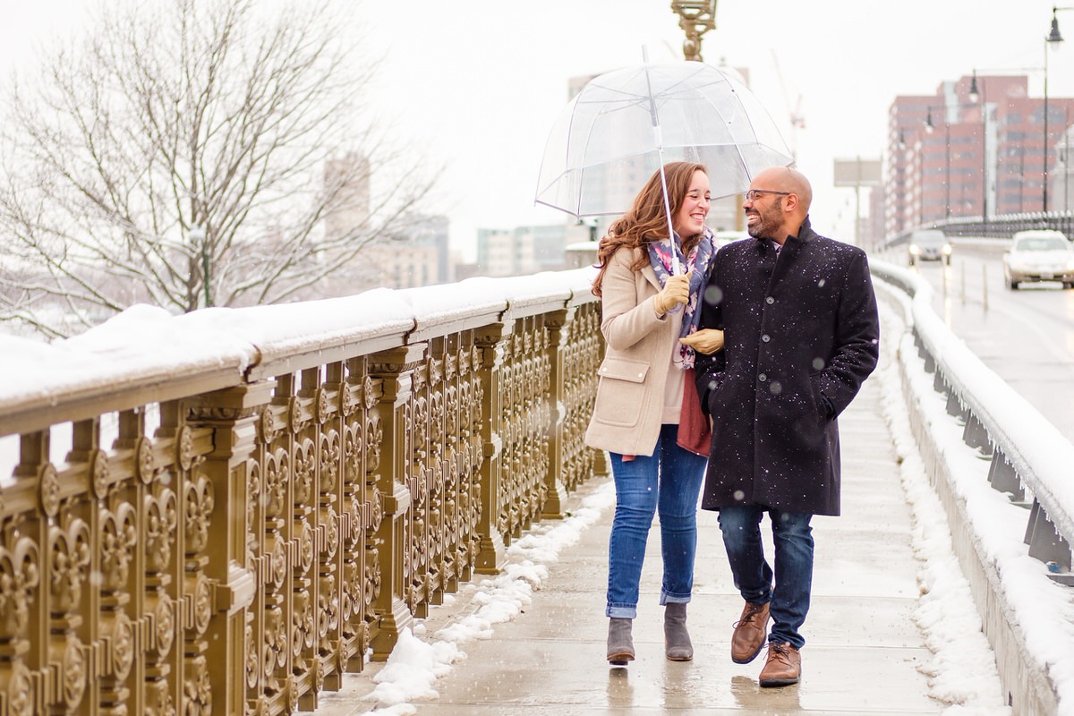 Engaged couple walks on the Longfellow Bridge with a clear umbrella on a snowy winter afternoon in Boston.