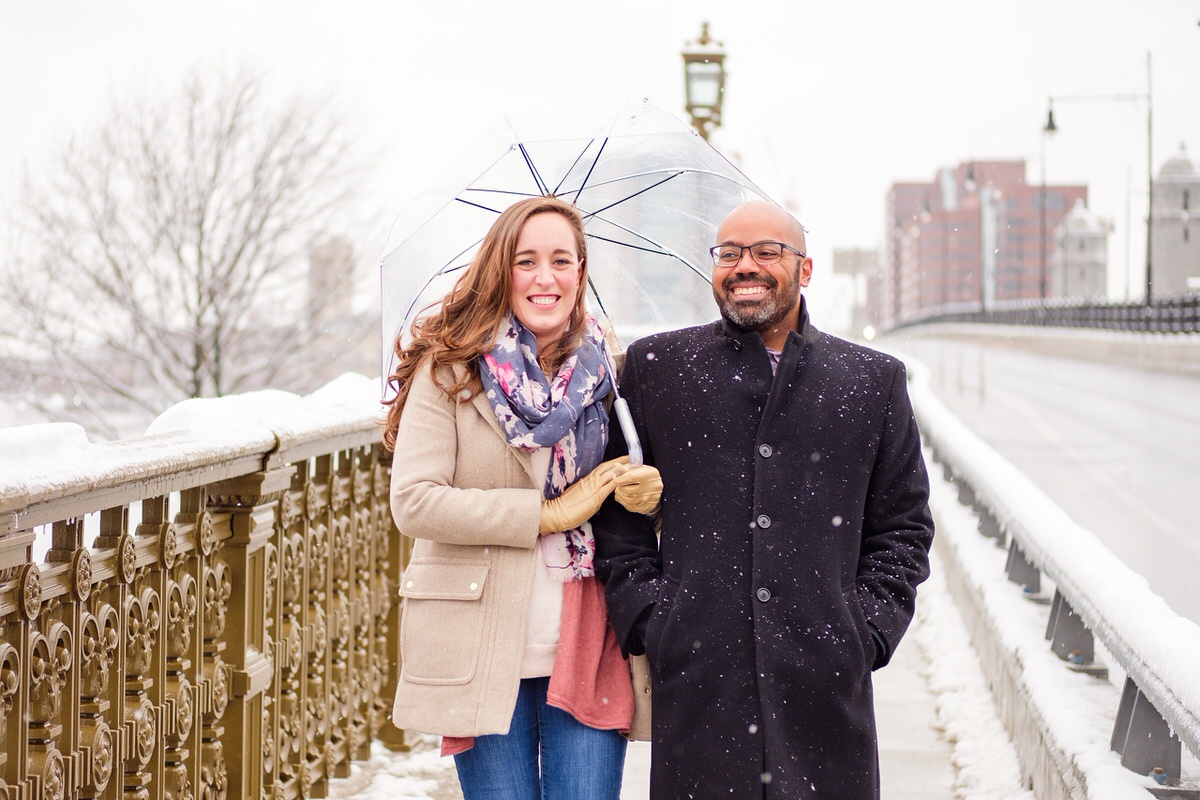 Engaged couple walks on the Longfellow Bridge in Boston with a clear umbrella on a snowy day.