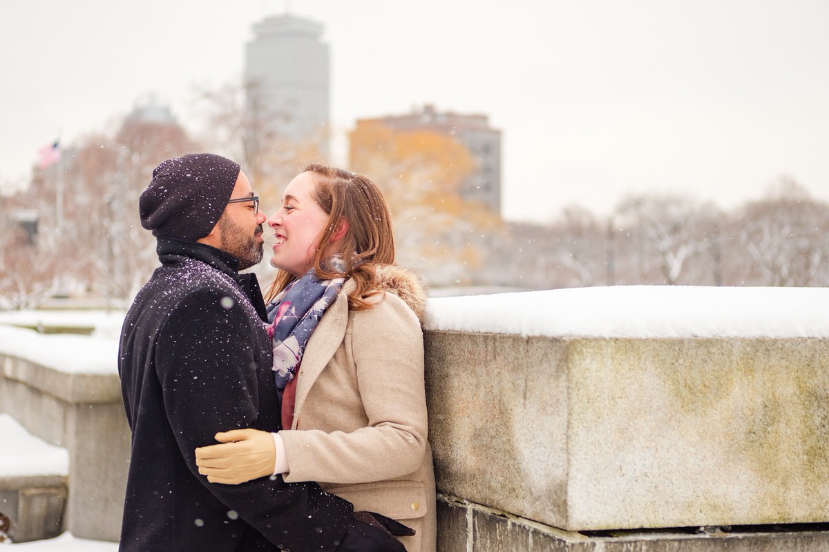 Engaged couple snuggles on a snowy winter day on the Charles River Esplanade in front of the Boston skyline