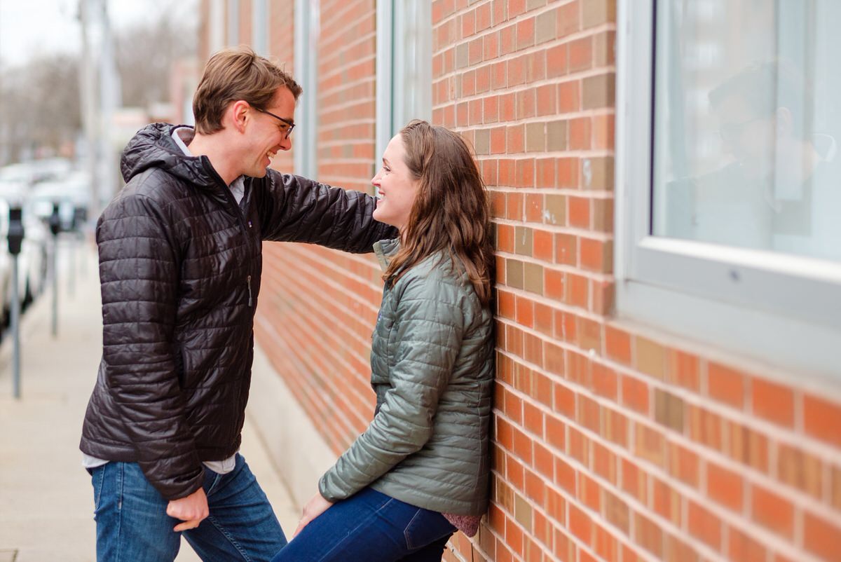 Engaged man leans against a brick wall with his fiance in Boston MA during an engagement photo shoot