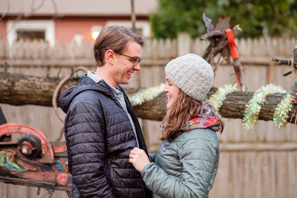 Adorable engaged couple in coats and a beanie hat pose by urban artwork on the Minuteman bike path in Davis Square