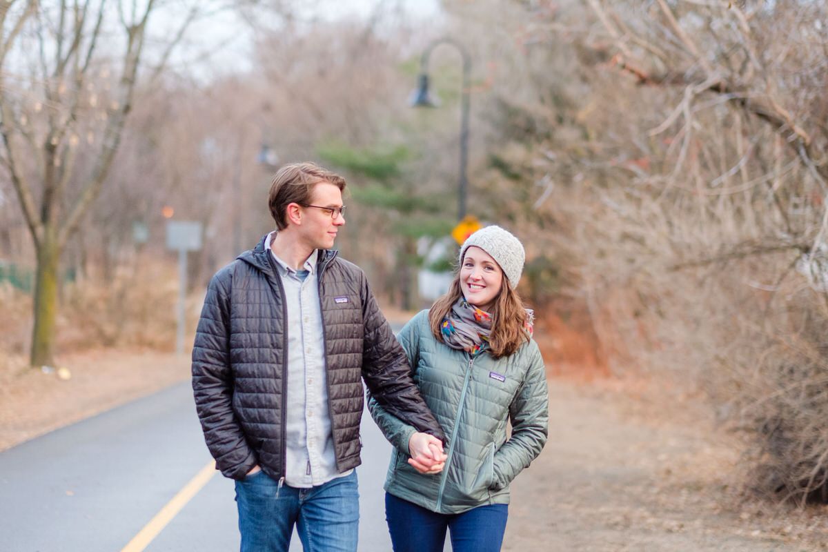 Engaged couple in Patagonia coats walks down the tree-lined Davis Square bike path during an engagement photo session