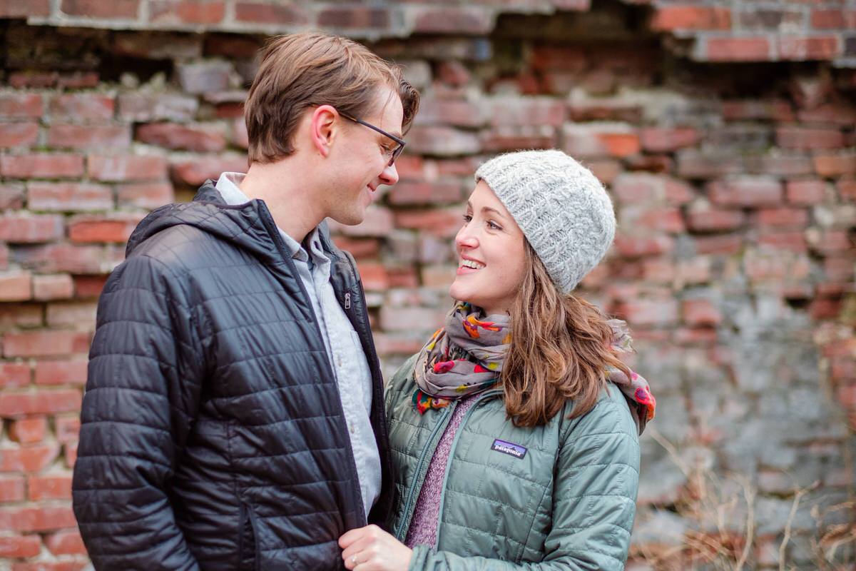 Engaged couple in winter coats poses in front of a brick wall in an alley in Somerville