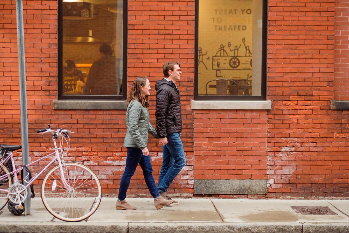 Couple in coats walk by a brick wall during a winter engagement session in Somerville, MA