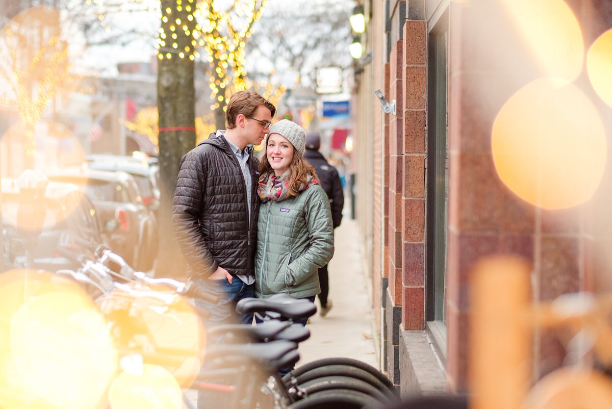 Adorable engaged couple in coats snuggles by twinkling holiday lights on the trees in Davis Square