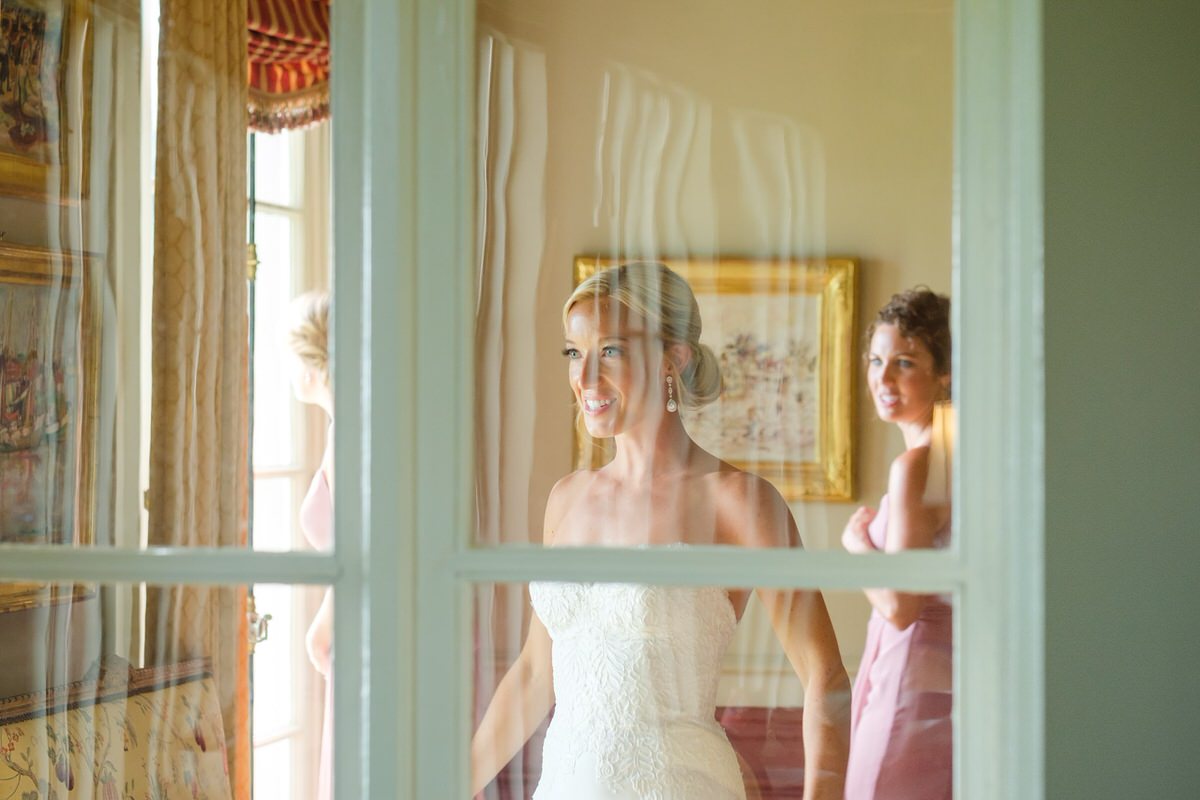 Blonde bride with an updo stands behind a glass window, looking at herself in a mirror as she gets ready for a New England manor wedding