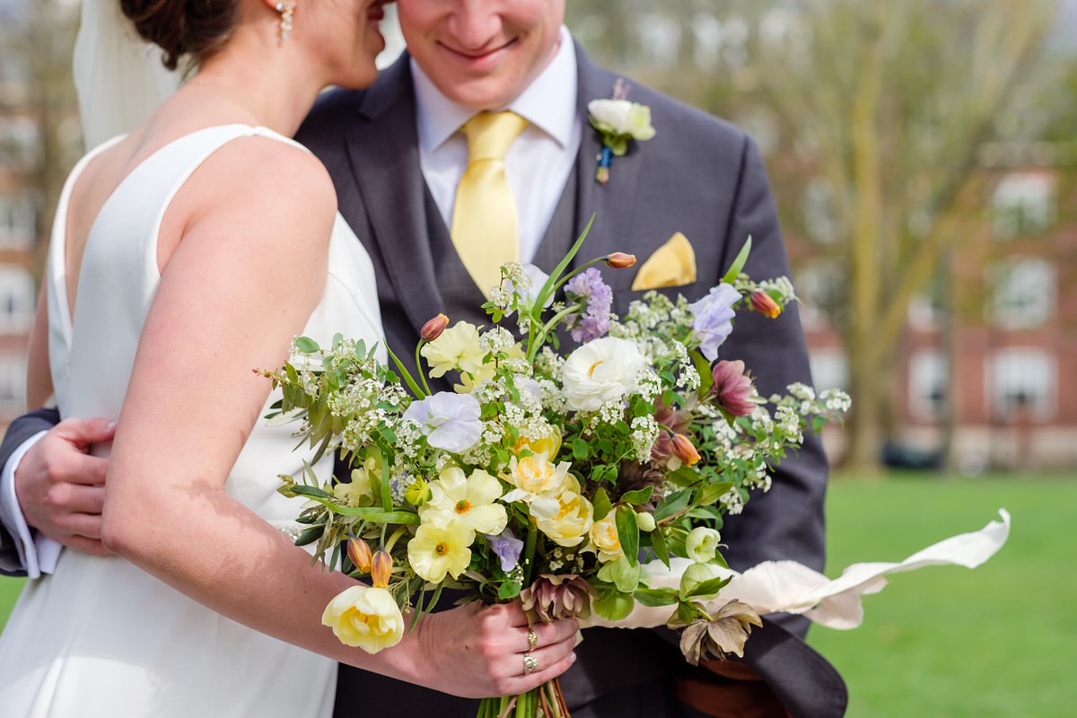 Bride and groom stand together before the wedding as she holds out a wild springy floral bouquet in yellow and purple