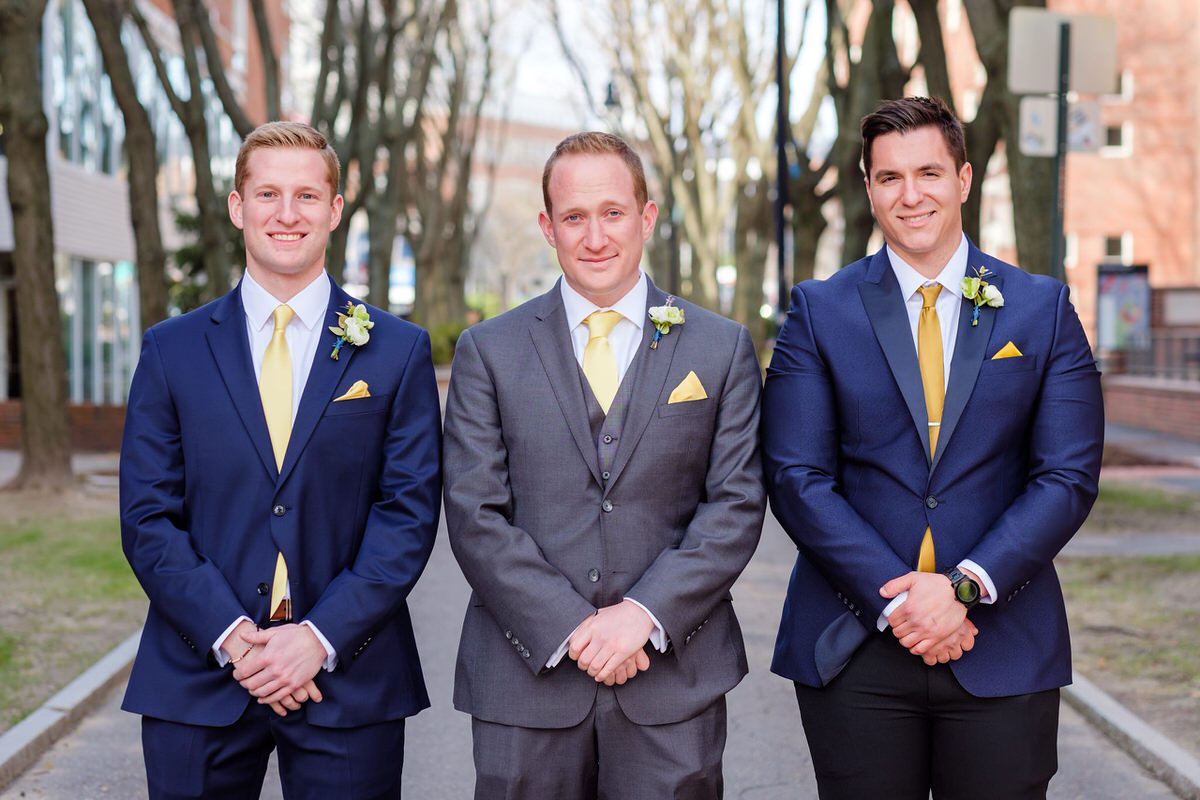 Groom in a grey suit poses formally with his two groomsmen in blue suits with yellow ties