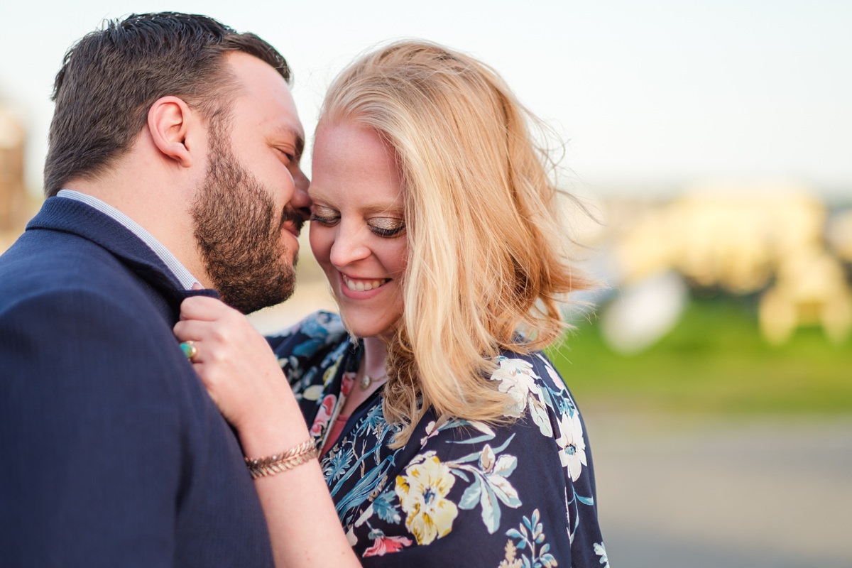 Blond woman in a floral navy shawl snuggles in front of the Salem historic waterfront with her fiance who is wearing a navy blue suit jacket