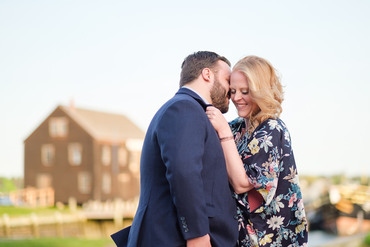 Blonde woman in a floral navy shawl snuggles in front of the Salem historic waterfront with her fiance who is wearing a navy blue suit jacket