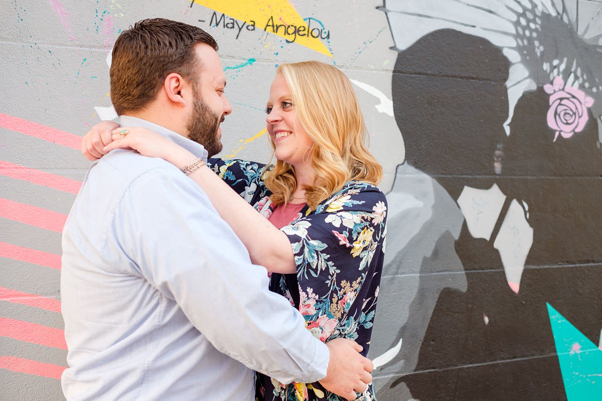 Blonde woman in a flowered shawl wraps her arms around her fiance in front of a mural at the Punto Art Museum in Salem, MA