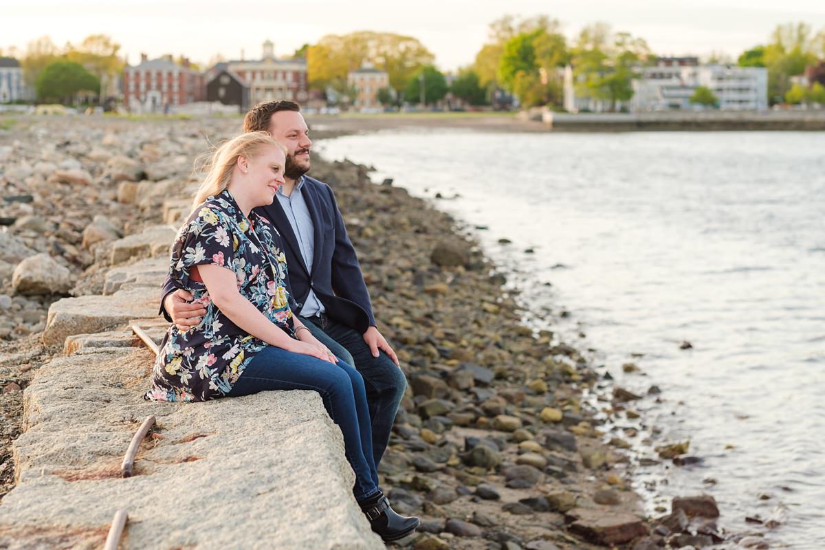 Man in a navy suit jacket holds his blonde fiance while they sit on a ledge overlooking the ocean