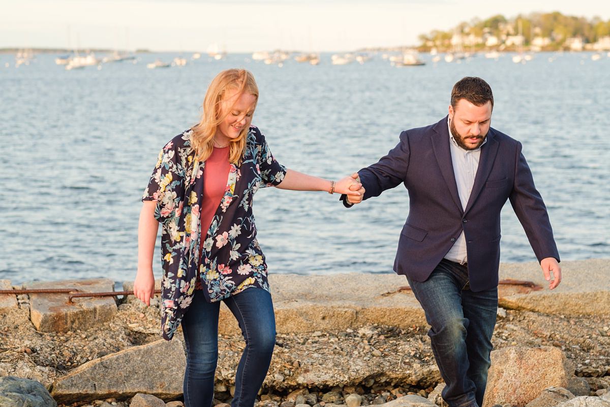 Man in a navy suit jacket holds hands with his blonde fiance as they climb over rocks near the ocean