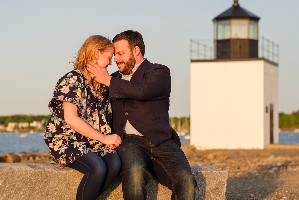 Man holds the face of his fiancee in front of the Derby Lighthouse in Salem while bathed in golden sunset light