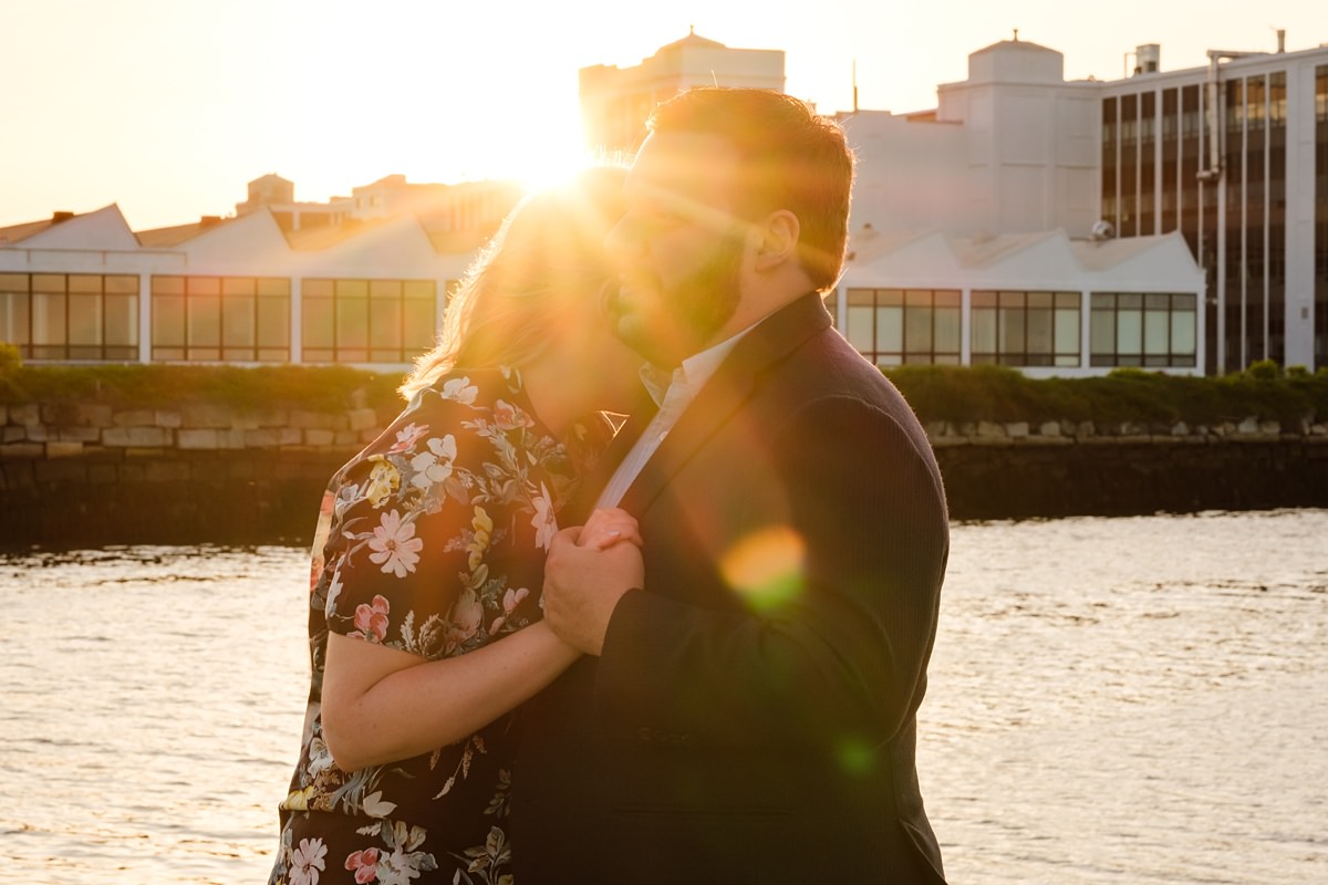 Man and woman hold each other as a golden sun flare shines on their heads in front of the water in Salem at sunset