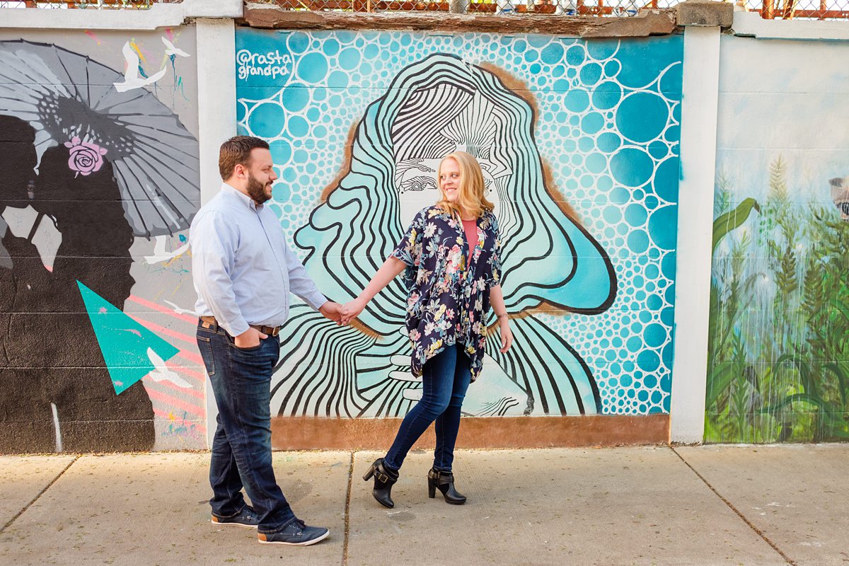 Engaged couple with woman in a flowered shawl holds hands while walking in front of a vibrant blue mural at the Punto Art Museum in Salem, MA