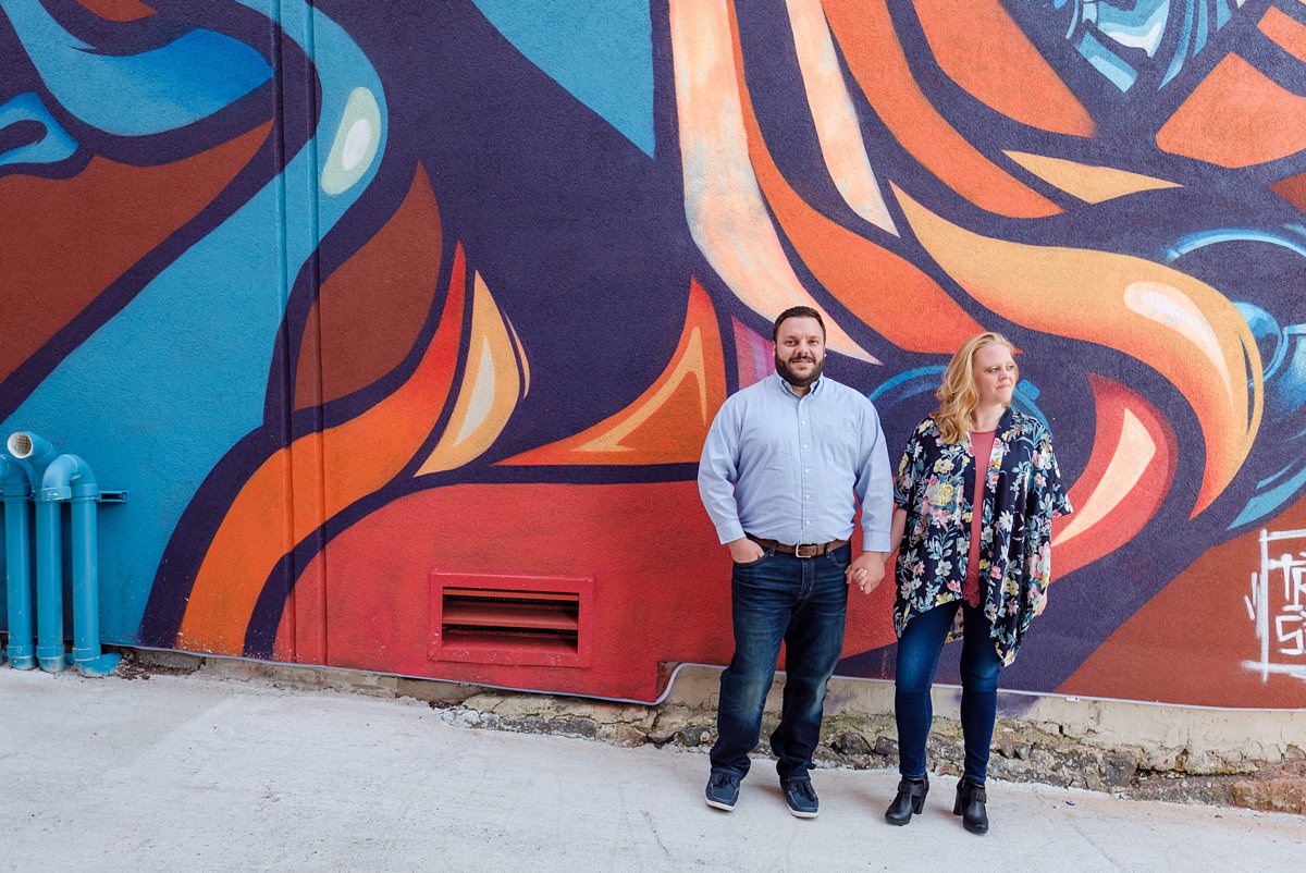 Woman in a floral shawl and man in a blue button down hold hands in front of a vibrant orange and blue mural.