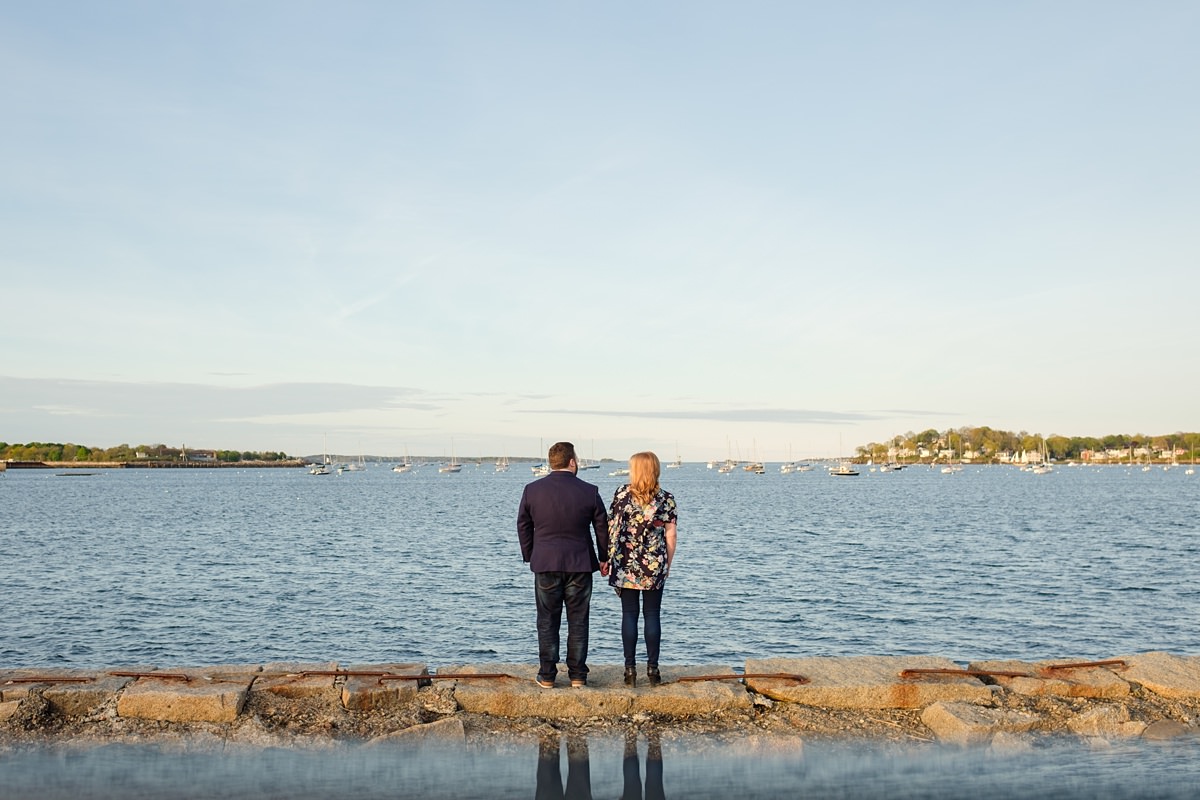 Man in a navy suit jacket holds hands with his blonde fiance while they stand on a ledge overlooking the ocean