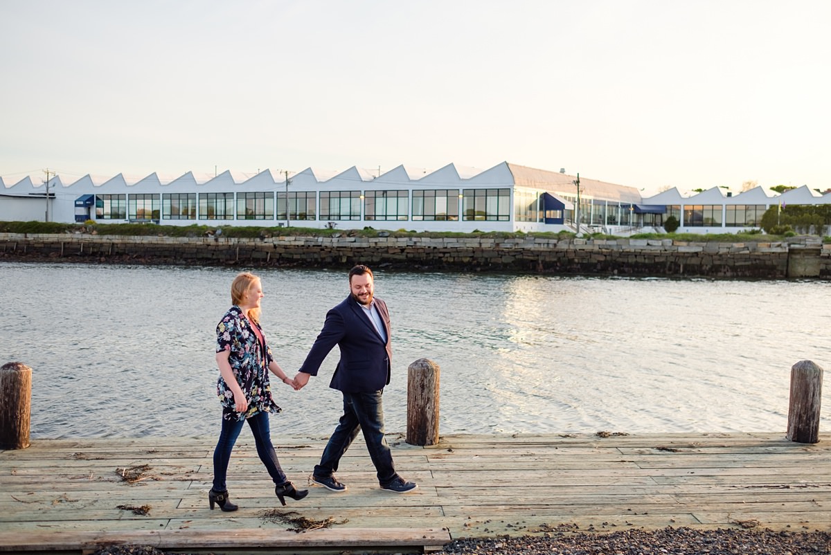 Man leads his fiance along an old wooden boardwalk as they stroll in front of the ocean in Salem, MA