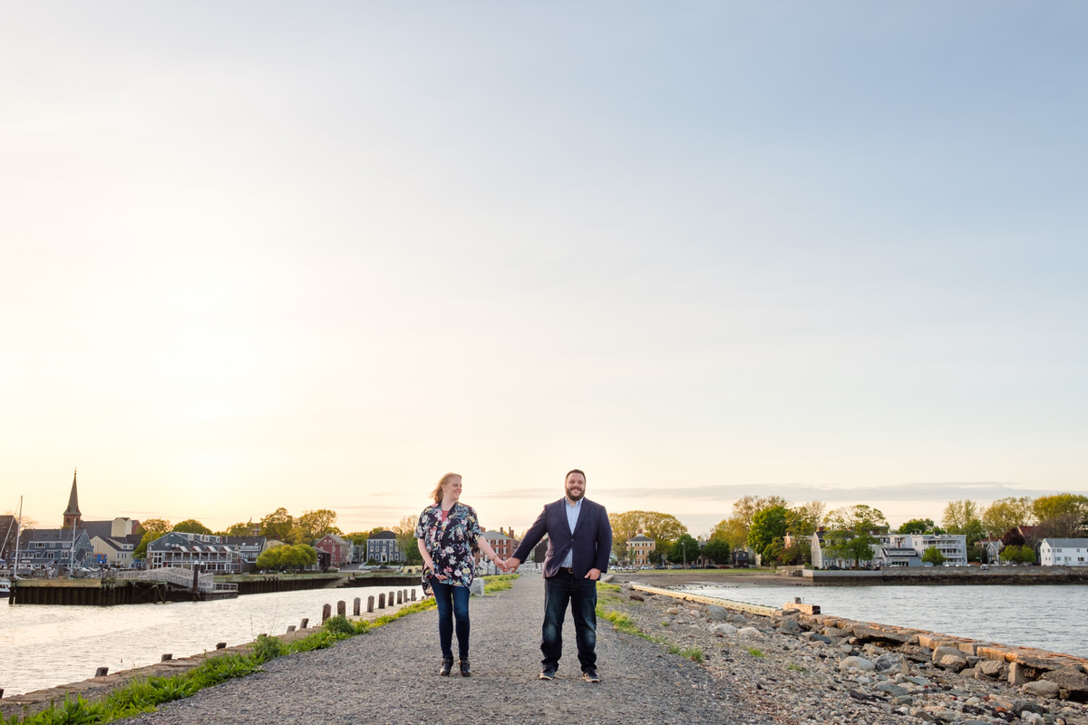 Engaged man and woman hold hands on the historic Salem waterfront Derby peninsula as the sun sets.