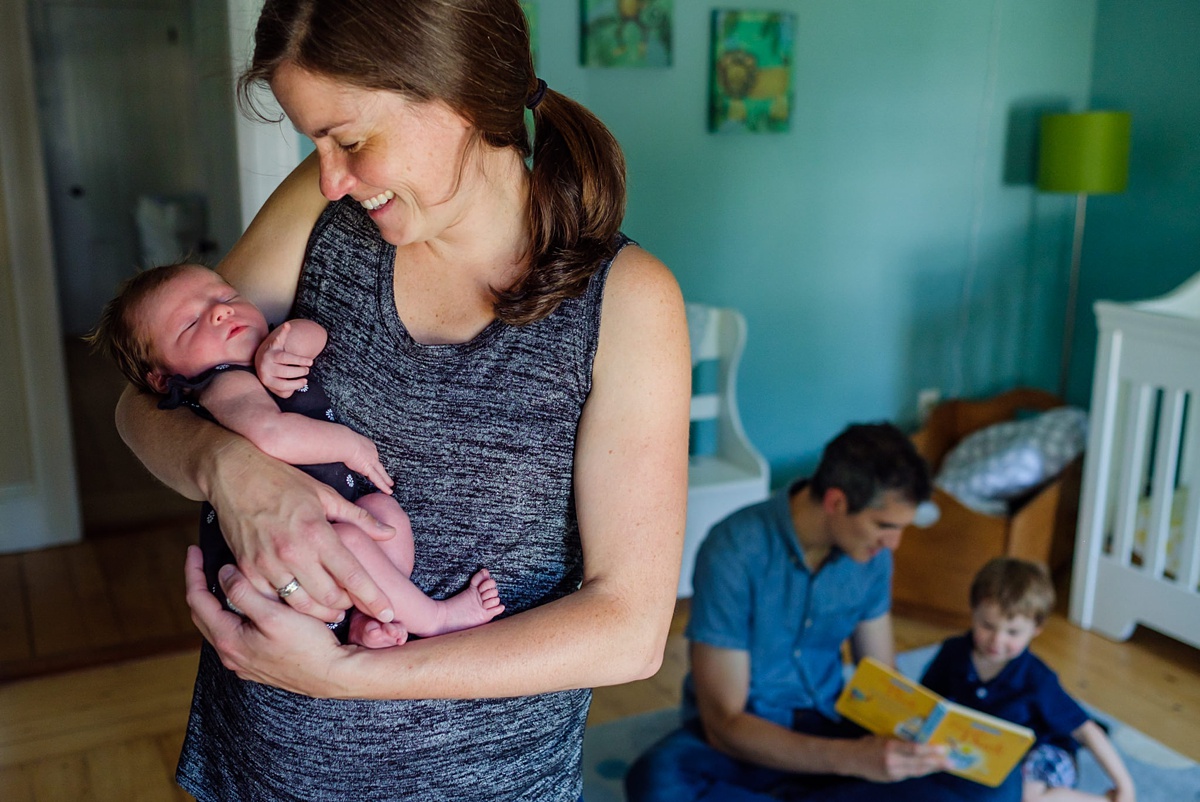 Mom holds her newborn baby girl while dad reads to their son in the background