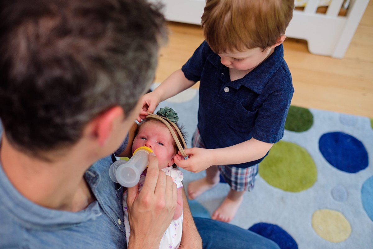 Dad feeds newborn daughter while toddler son puts a headband on her