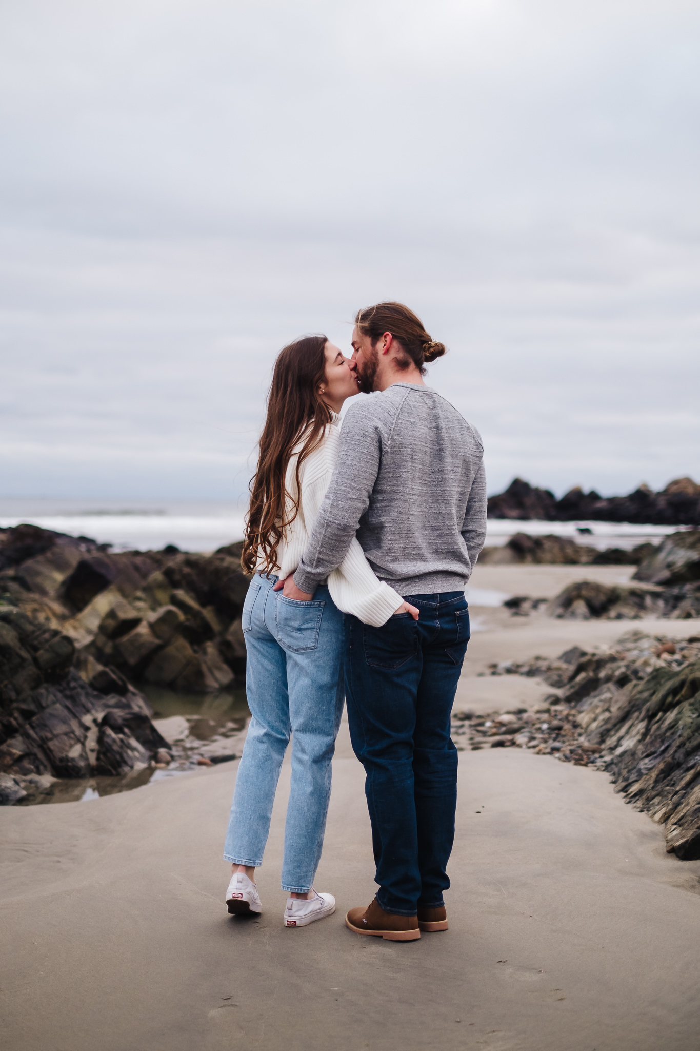 White couple in jeans and sweaters kisses with their hands in each other's back pockets at Cape Hedge Beach in Rockport