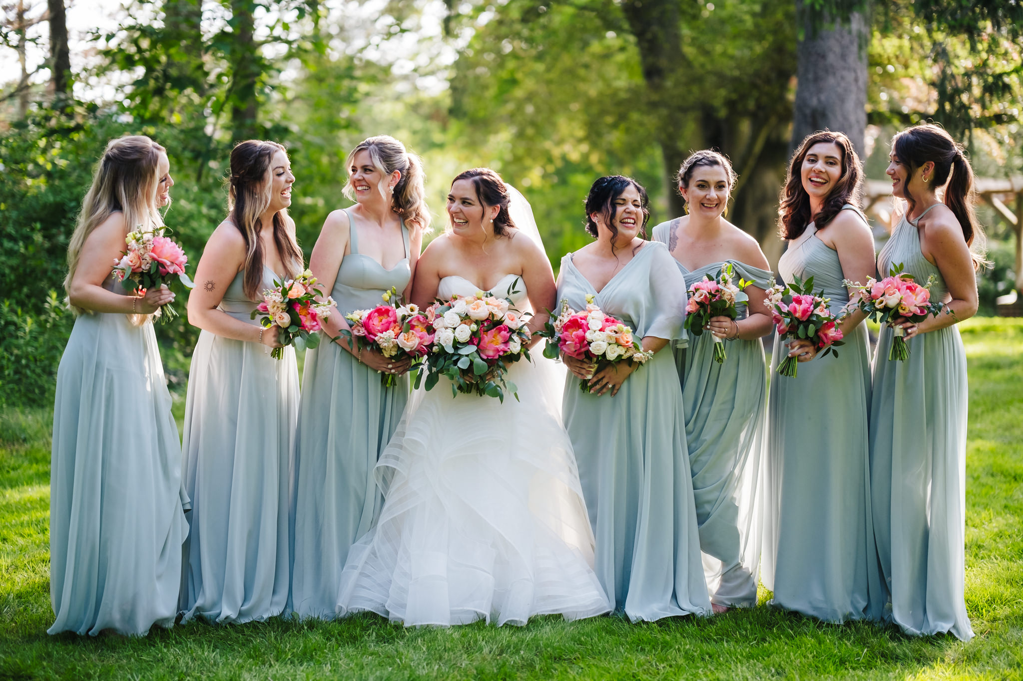 Bride and bridesmaids in minty sage dresses smile at each other in a wooded area