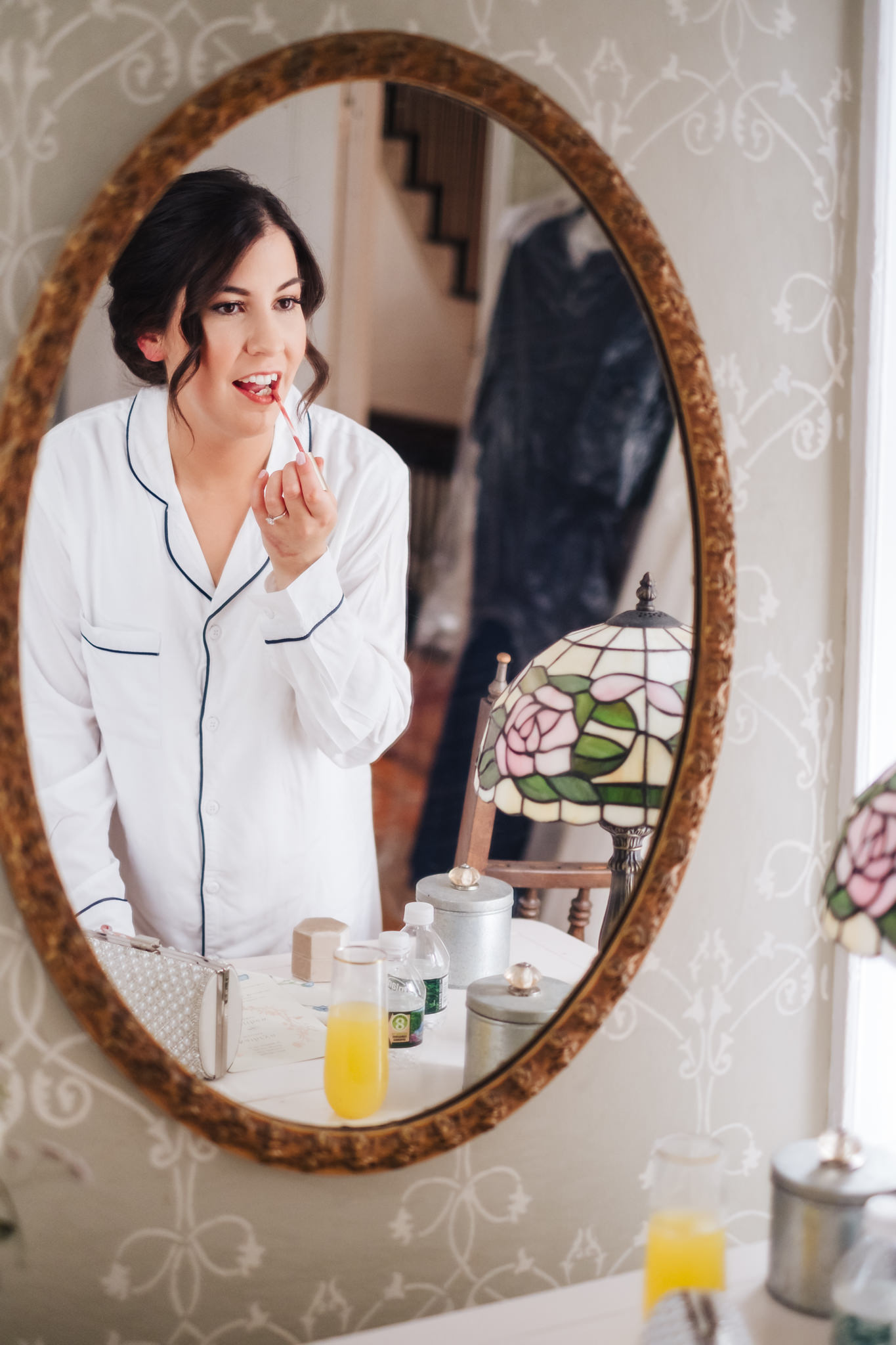 Bride applies red lip gloss while looking in an ornate oval mirror