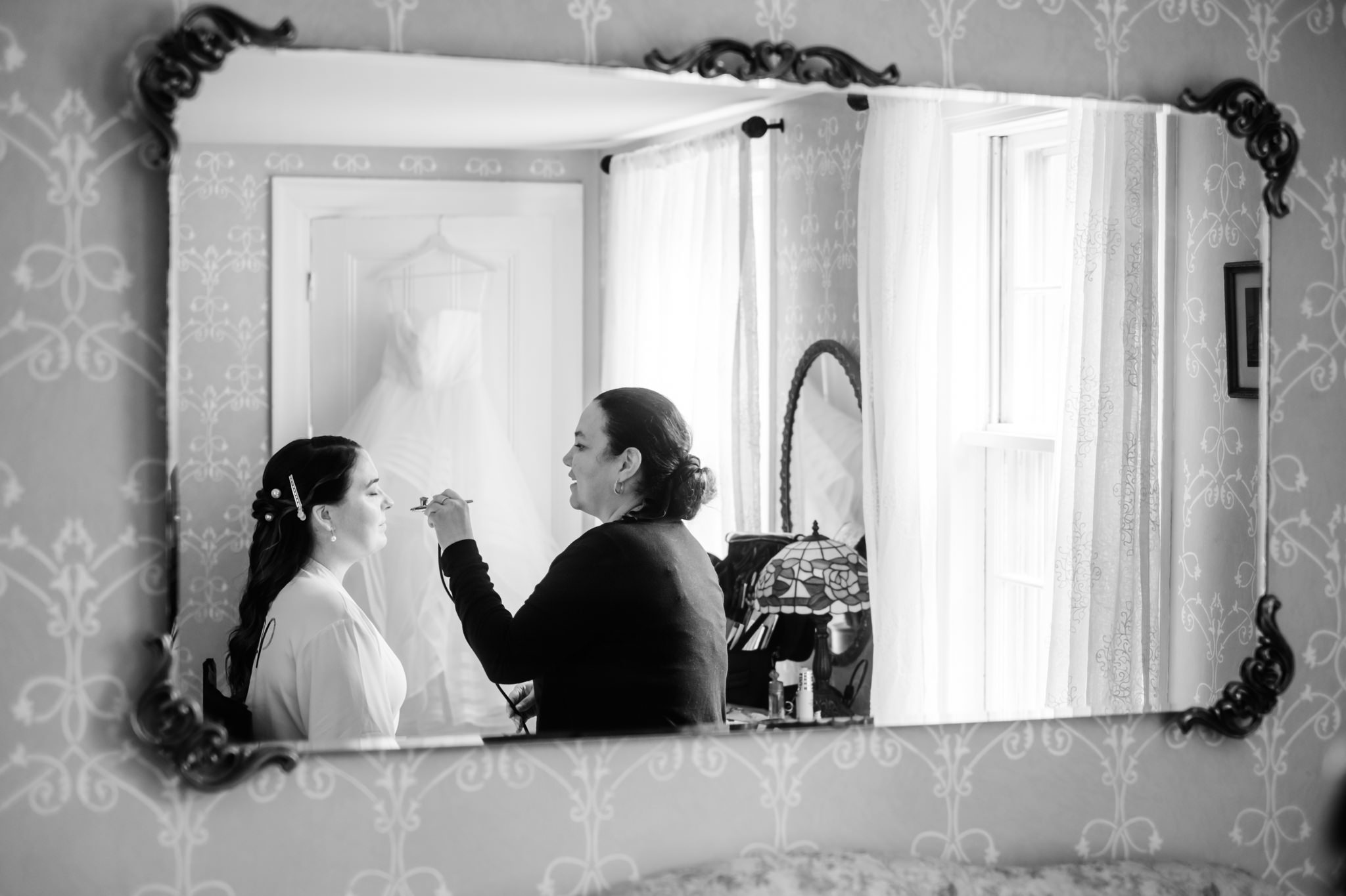 Through a mirror, a woman in black applies airbrush makeup to a bride in a white robe