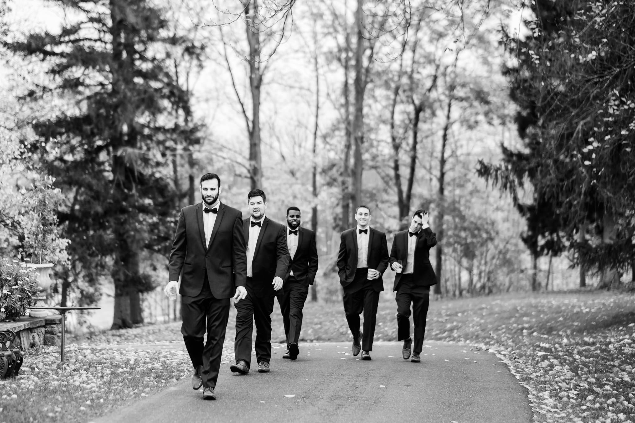 5 groomsmen in tuxes and bowties walk forward on a road